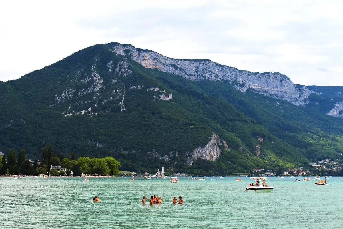 A tranquil lake scene with swimmers and boats against a majestic mountain backdrop, perfect for a summer retreat.